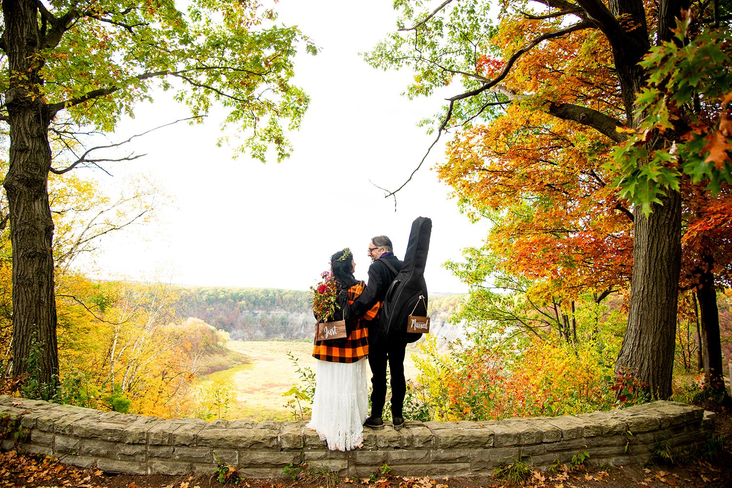 Two figures stand together on a stone outlook point surrounded by vibrant autumn foliage in a scenic natural setting.