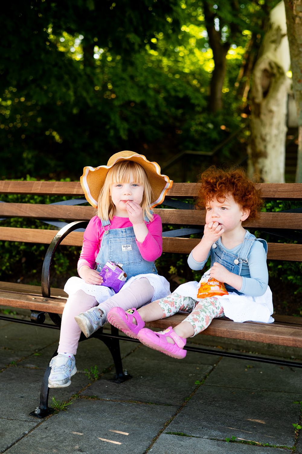 Two young children in denim overalls sitting on a park bench with trees in the background.