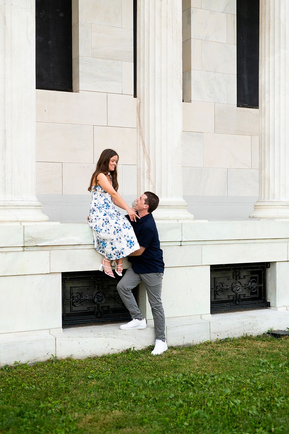 A cheerful couple poses playfully against white stone columns on a grassy lawn outside a neoclassical building.