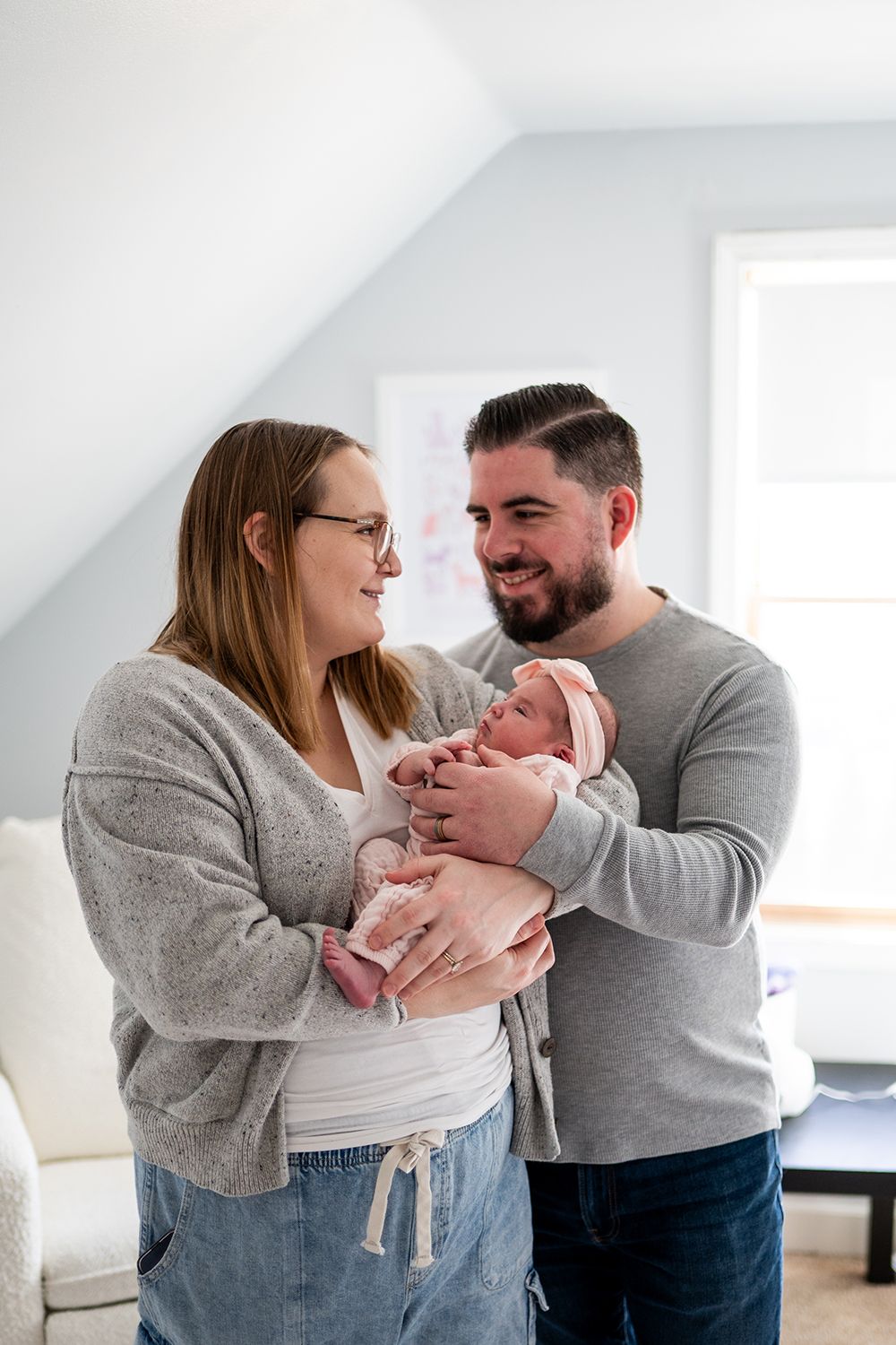 New parents share tender moments with their newborn baby while dressed in cozy gray sweaters in a bright white room.
