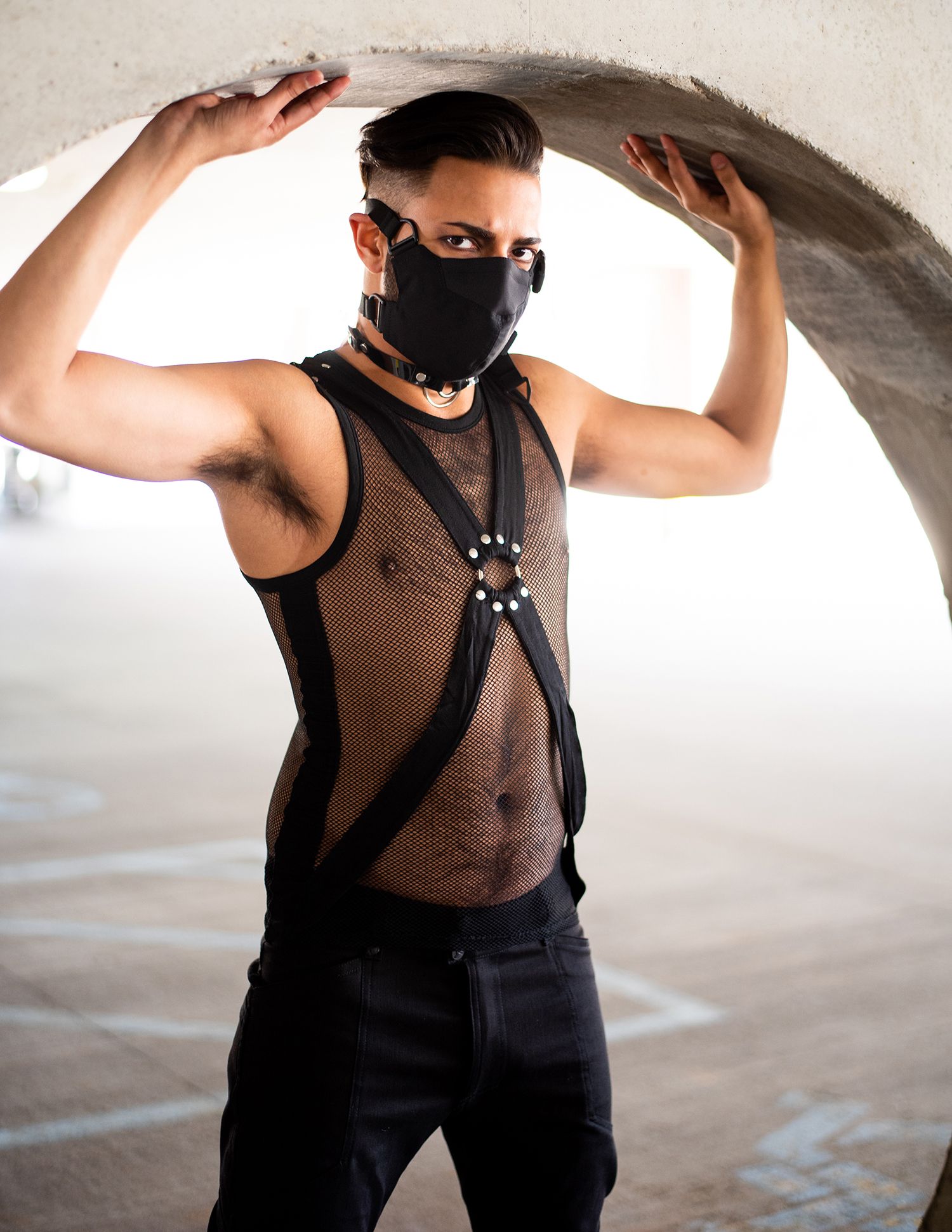 Person wearing black mask and harness poses with arms raised under curved archway in artistic fashion portrait.