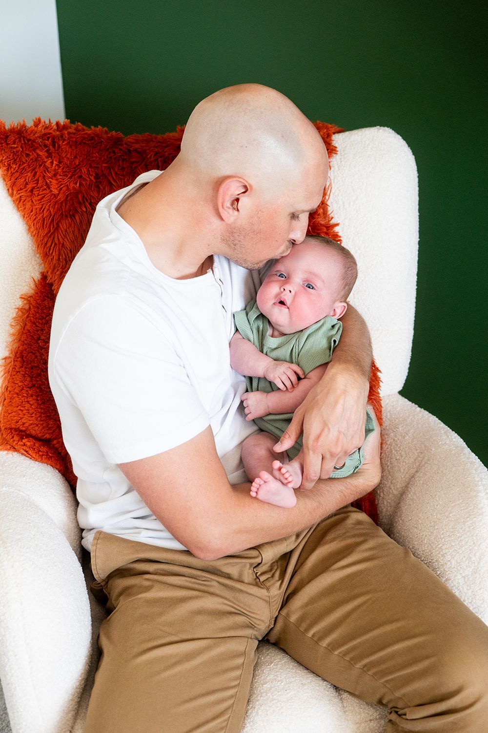 Intimate family moment in white armchair with reddish orange pillow and mint green baby outfit.