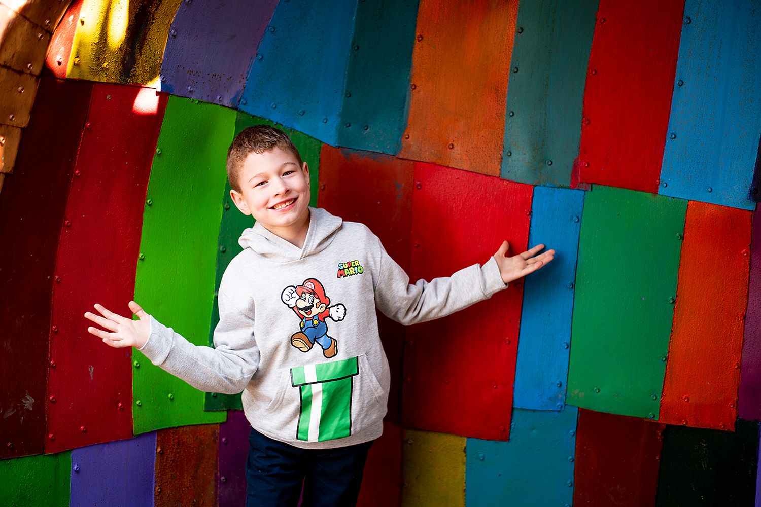 Energetic pose against a multicolored striped wall background while wearing a Super Mario graphic sweater.