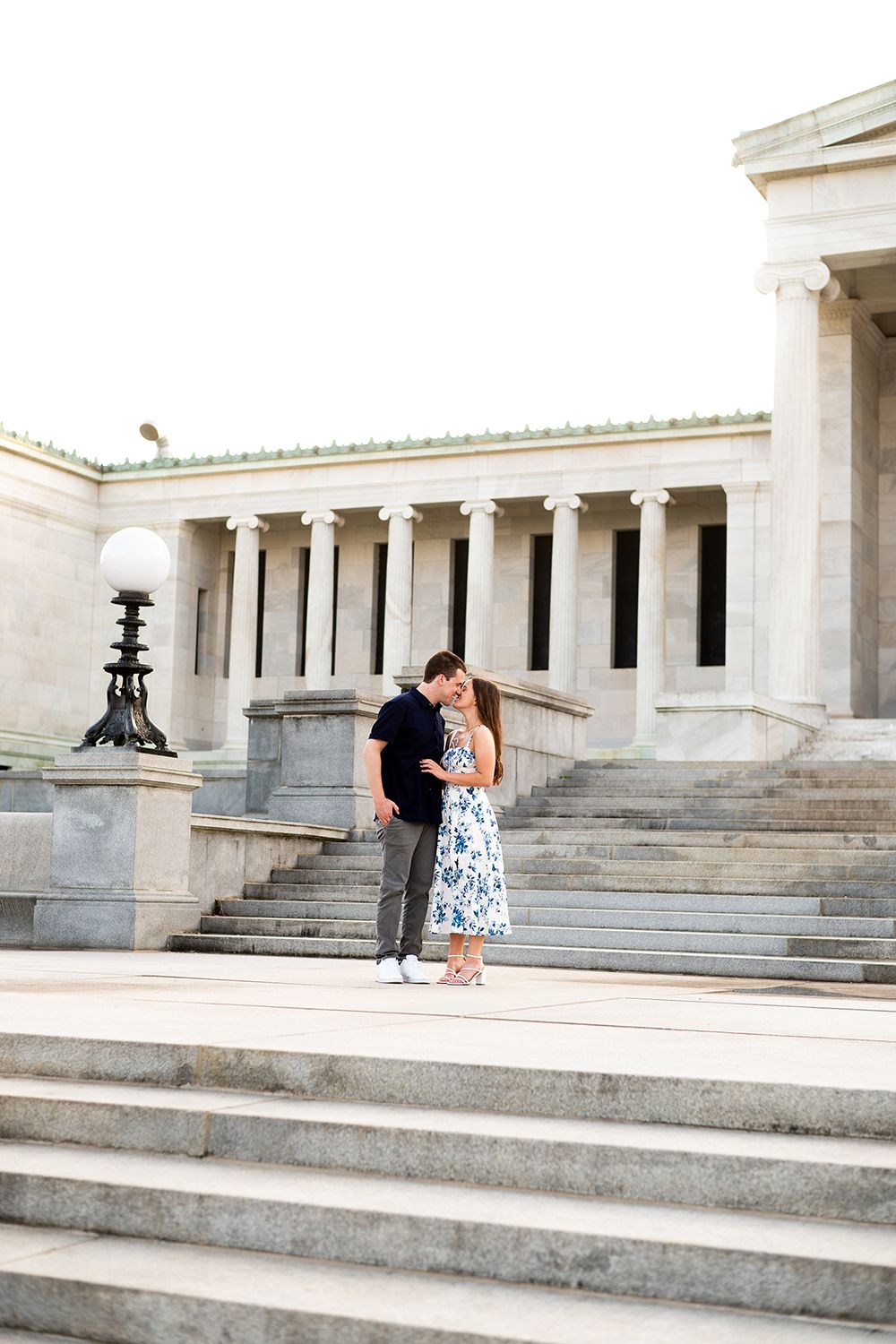 A romantic couple embraces on grand marble steps with elegant columns and architecture in the background during sunset.