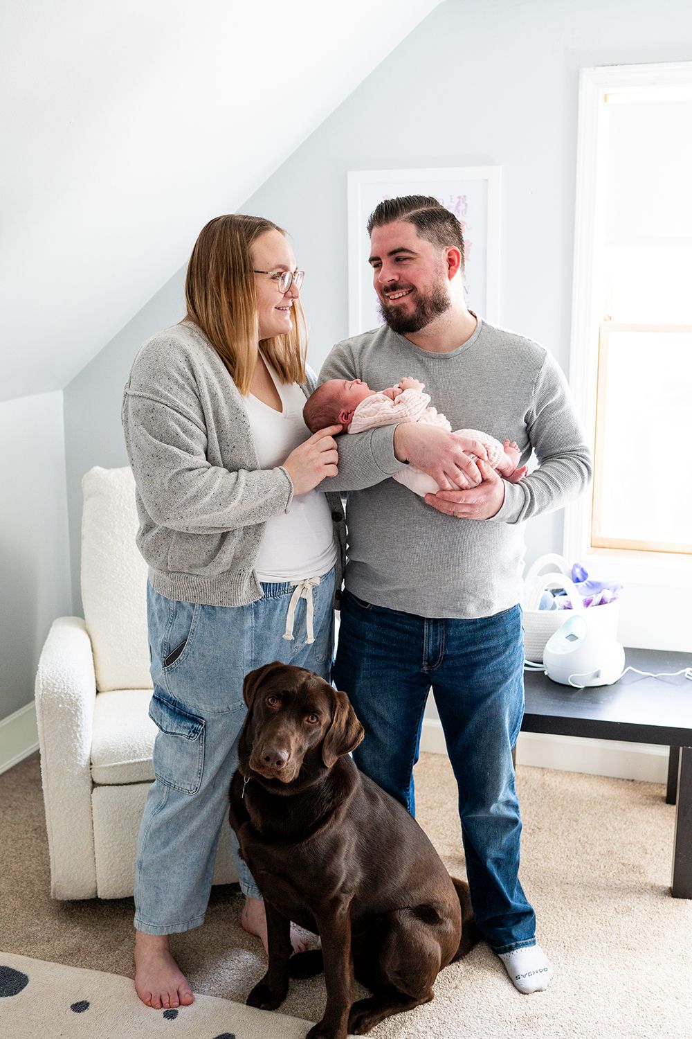 A couple holds their newborn baby while their chocolate Labrador dog sits attentively beside them in a bright living room.