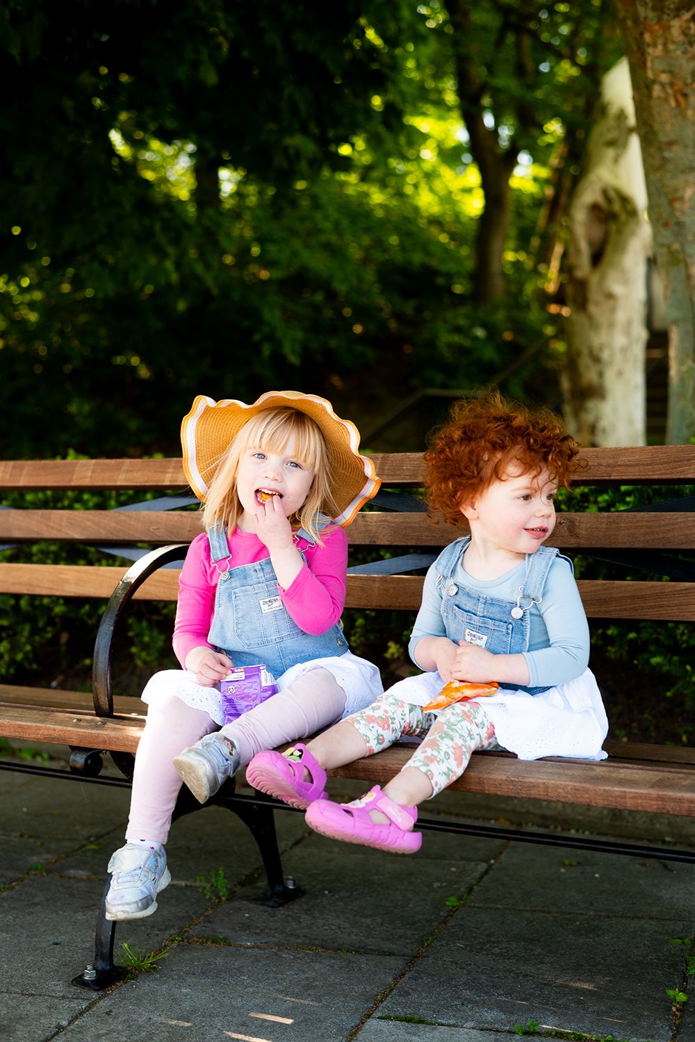 Two children making silly faces while sharing snacks on a wooden bench in a park setting.