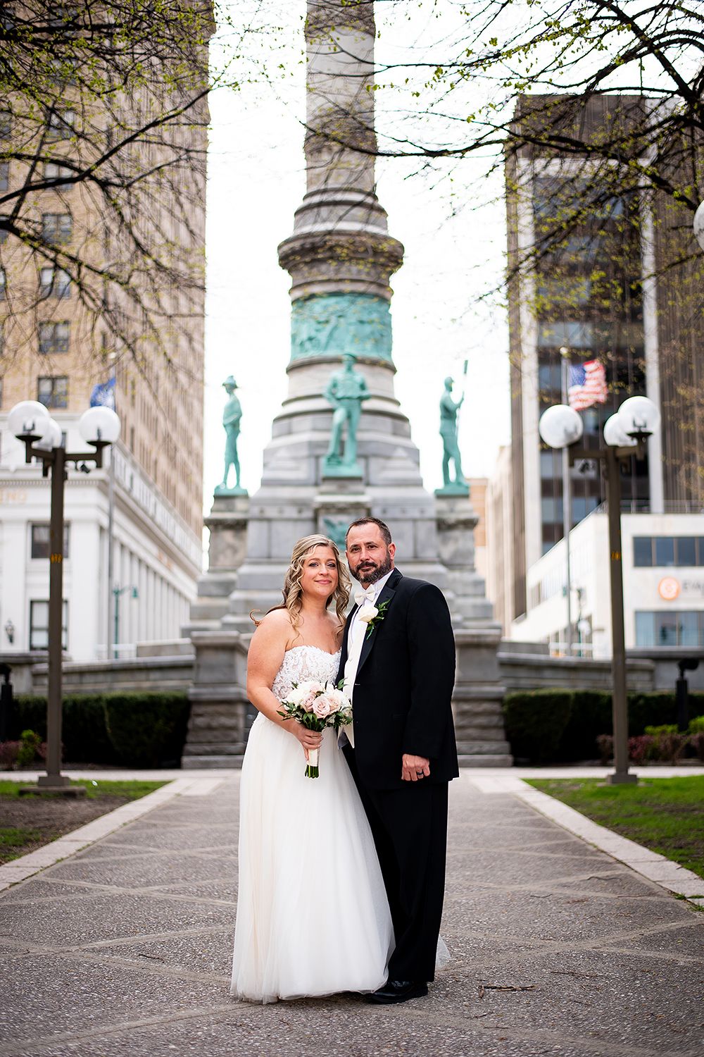 A couple poses for a wedding photo in front of a grand monument with ornate columns and statues in an urban setting.