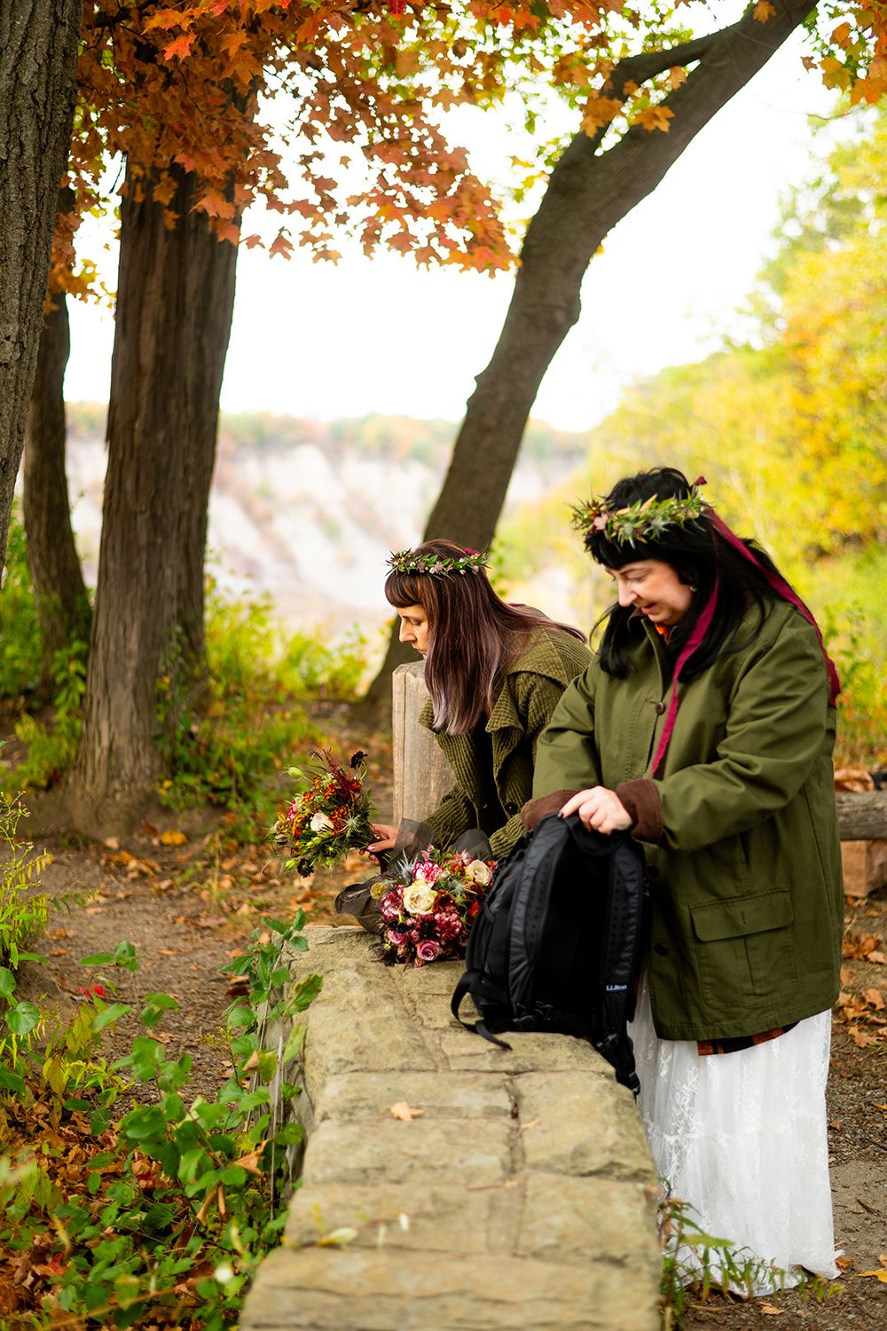 Two people wearing floral crowns and autumn coats explore a stone path surrounded by fall foliage in golden light.