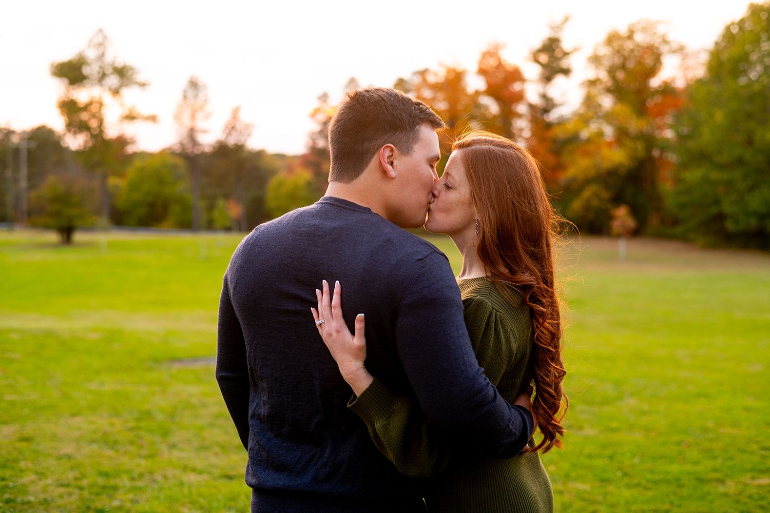 A couple sharing a romantic kiss in a park at sunset with autumn trees in the background.