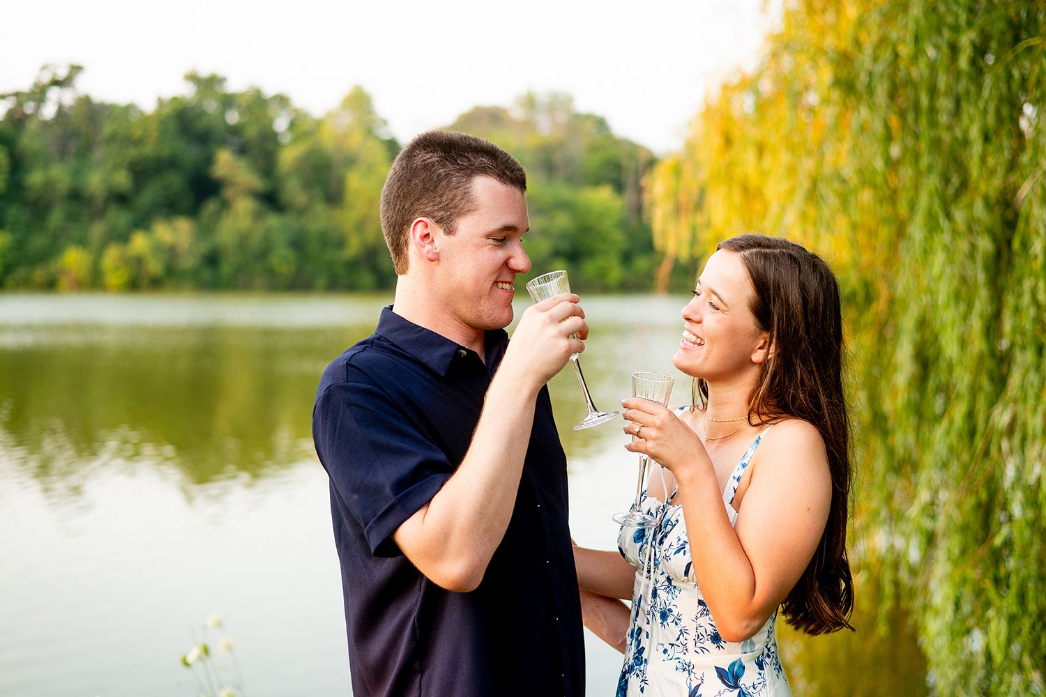 A romantic couple shares tender moments by a tranquil lake with weeping willow trees during a golden sunset photoshoot.