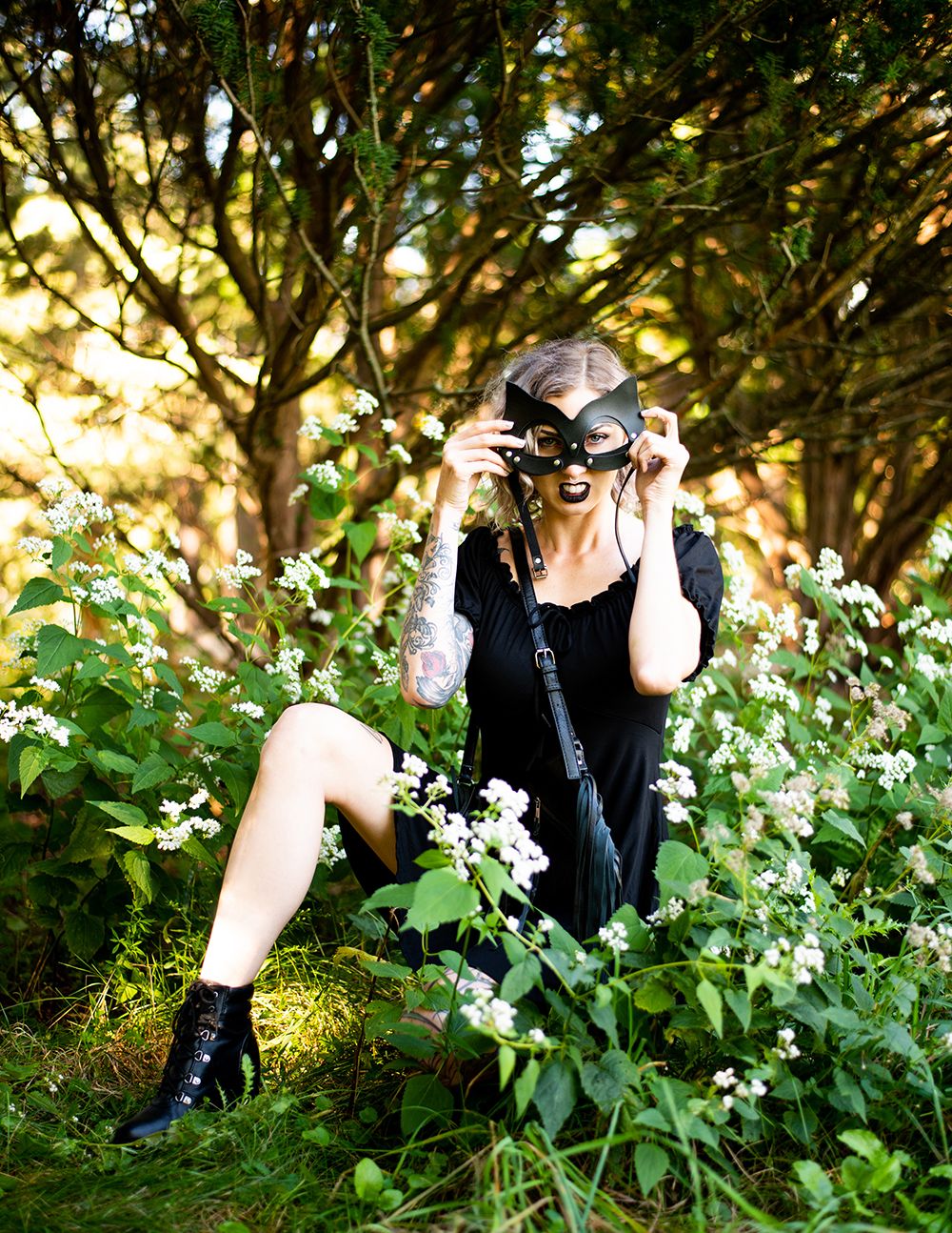 Photographer in black dress and boots crouches in garden taking photos among white flowers during golden hour.