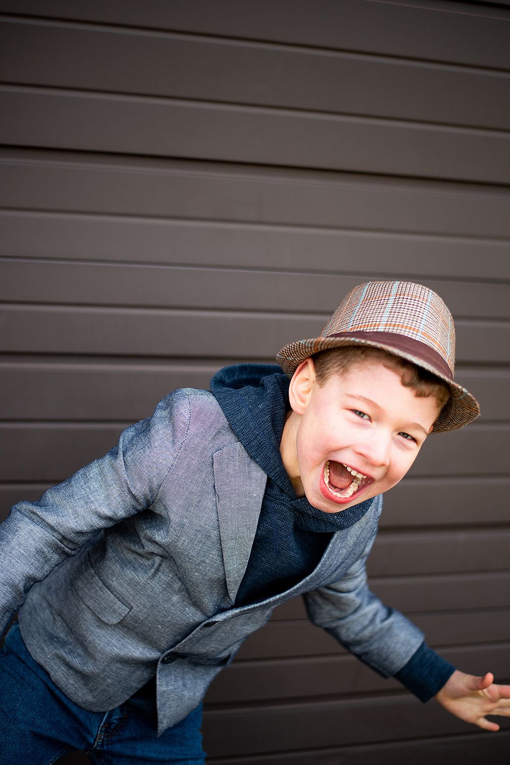 Young person in a plaid hat and gray jacket laughs joyfully against dark garage door background.