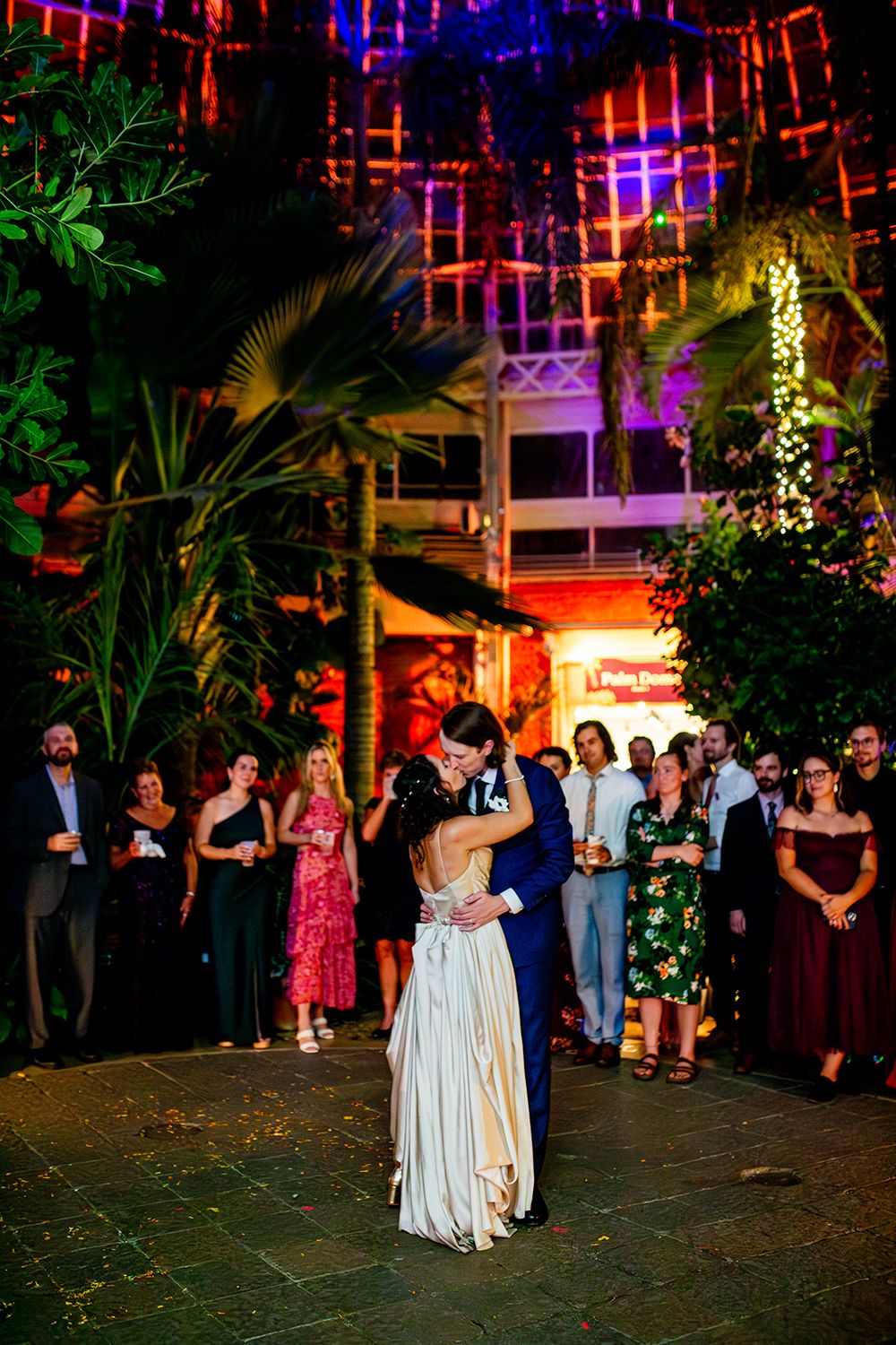 Guests dance and celebrate at an evening wedding reception under colorful lights in a lush tropical garden setting.