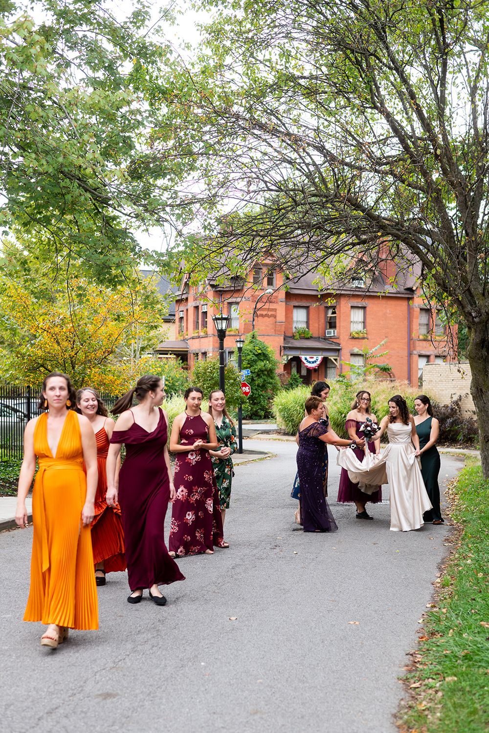 Wedding party walking down a scenic path with autumn trees and historic brick building in background.