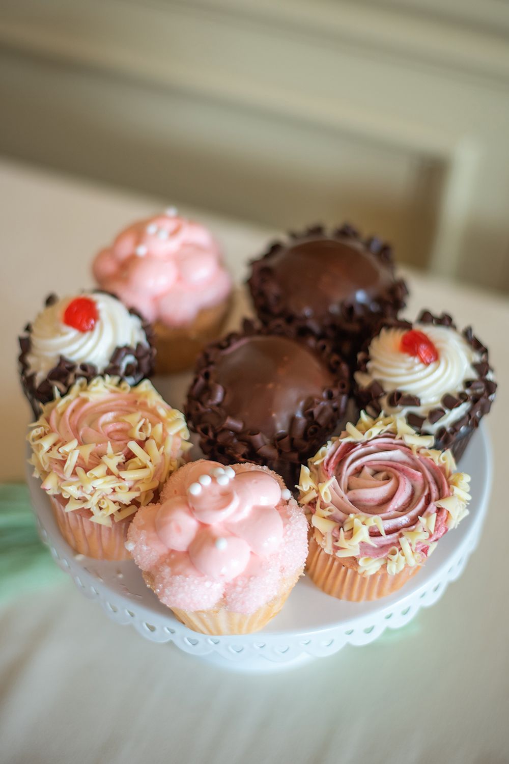Close-up of gourmet cupcakes featuring pink swirls, chocolate coating, and cherry decorations on a lace-trimmed stand.