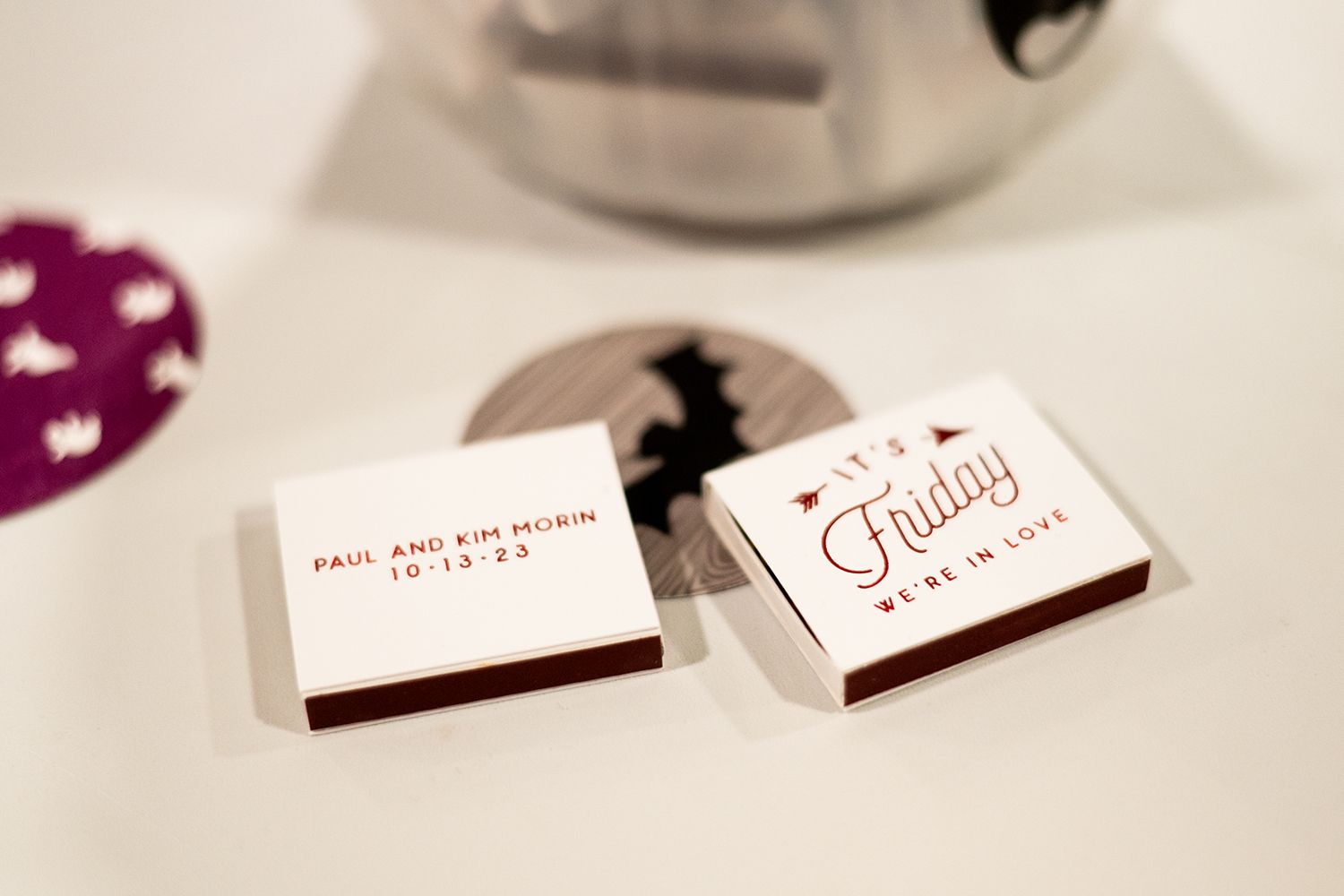 White matchboxes with red lettering that appear to be wedding favors sit on a table near a pink decorative item.