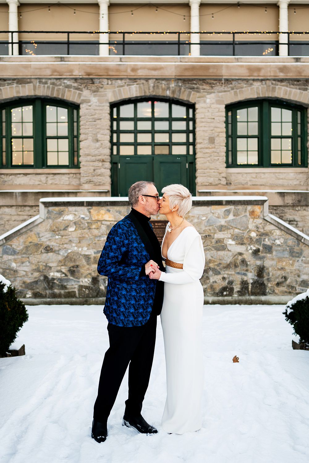 Two figures embrace in a romantic moment against a snowy backdrop with a stone building in the background.