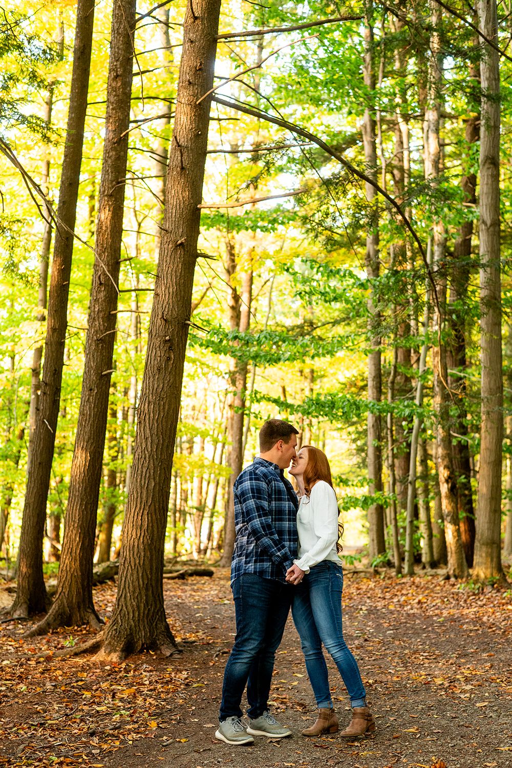 A romantic couple shares a tender moment on a woodland trail surrounded by tall autumn trees and golden light.