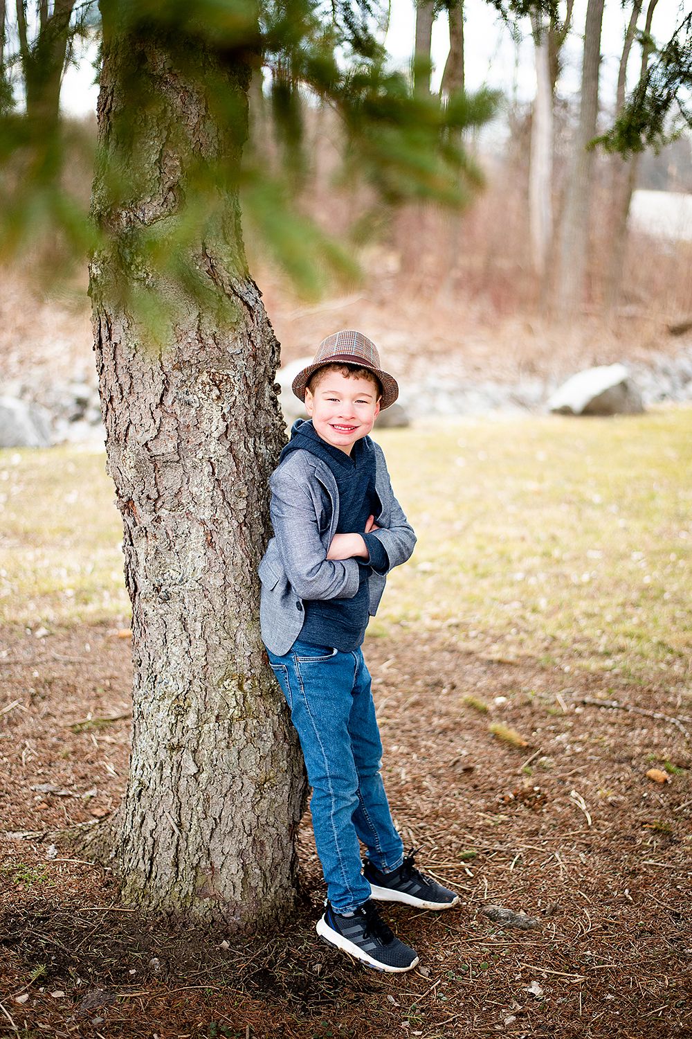 A young person wearing a brown hat and blue jeans leans against a large tree trunk in an outdoor park setting.