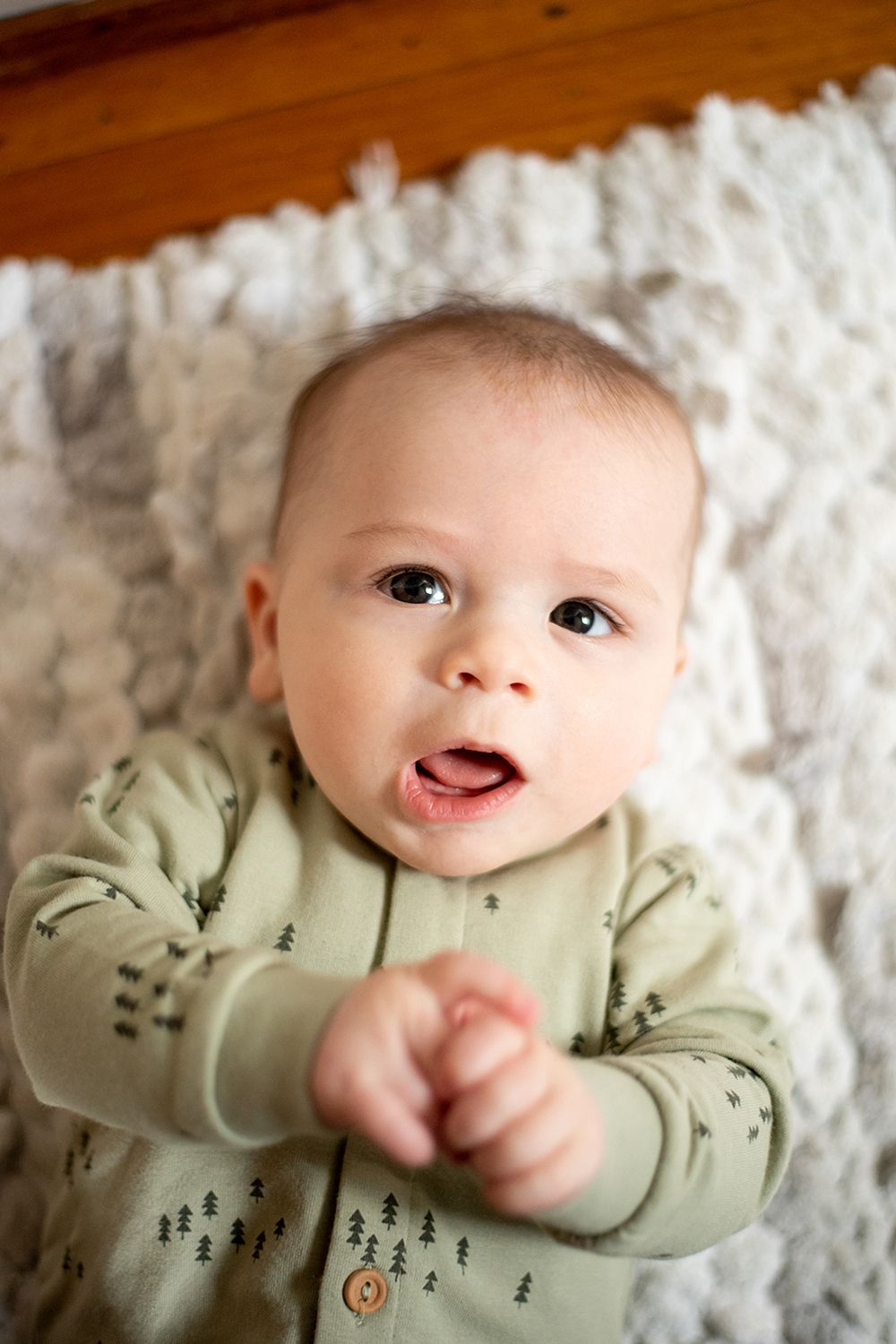 A small baby wearing a light green outfit with tree patterns lies on a white textured blanket.