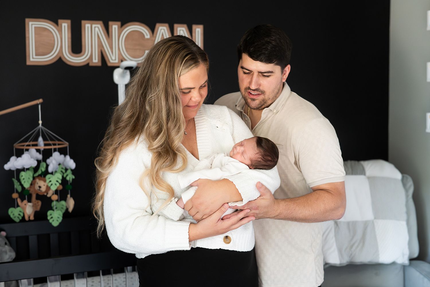 Parents tenderly hold their sleeping newborn baby in a peaceful nursery setting with a mobile hanging overhead.