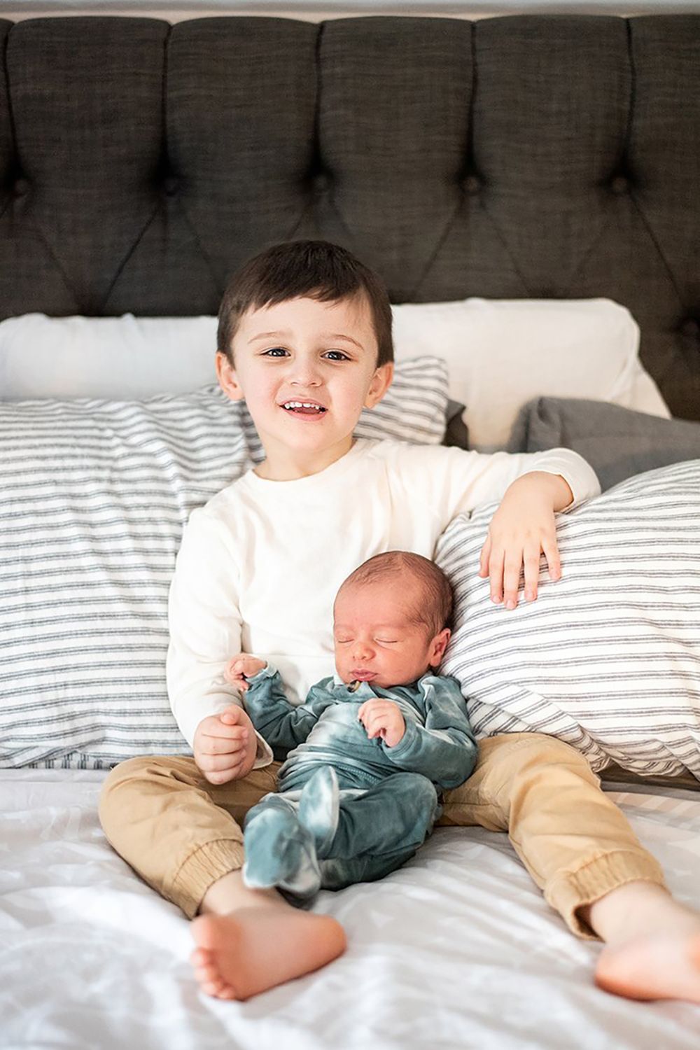 Two children sitting on a bed with striped bedding against a tufted grey headboard in natural light.