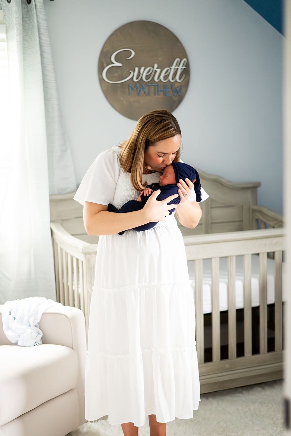 A woman in a white dress stands in a blue and gray nursery holding a baby near a white crib and round wall sign.