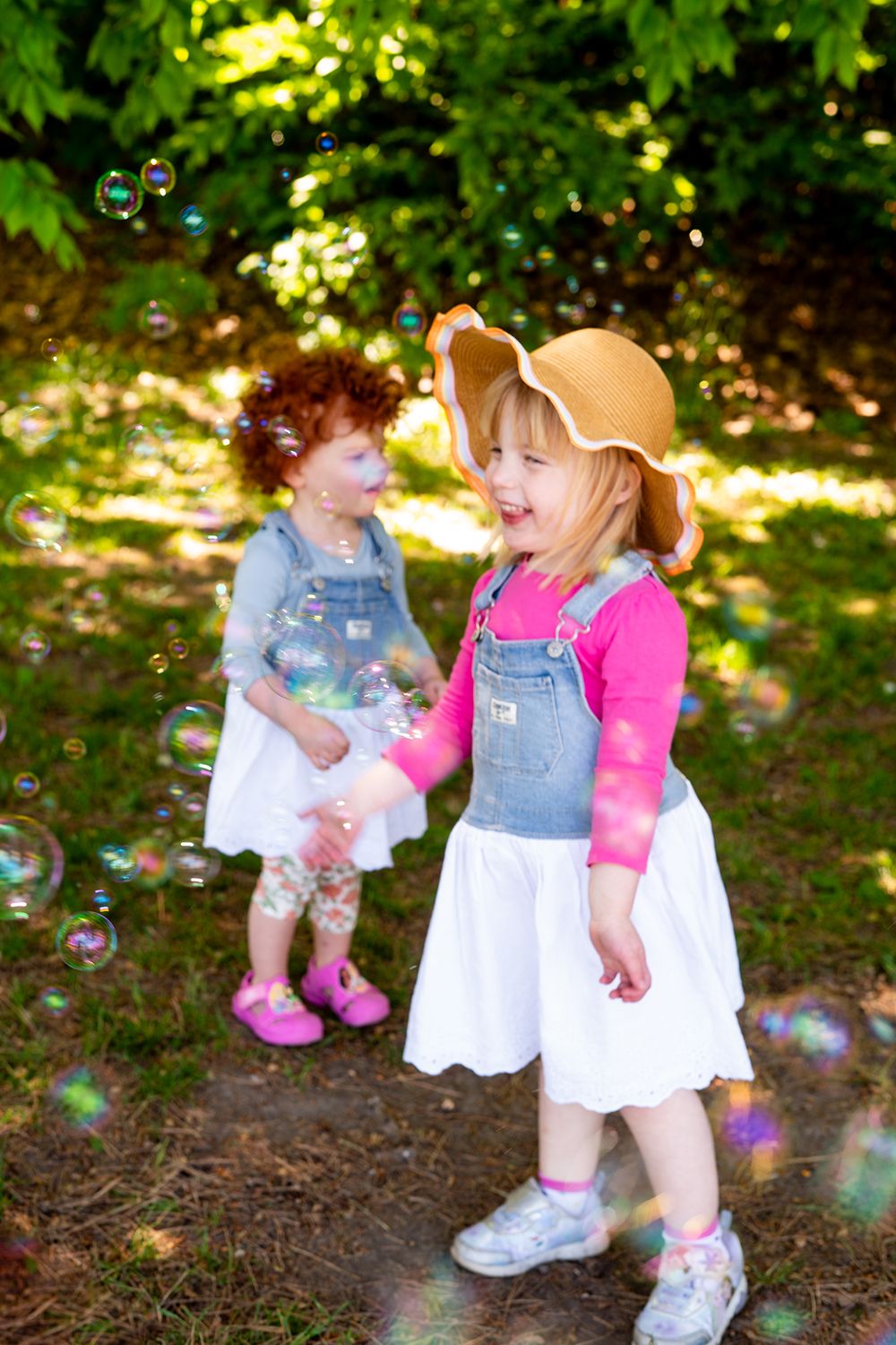 Two toddlers in denim overalls and white dresses playing with bubbles on a sunny day in a garden setting.
