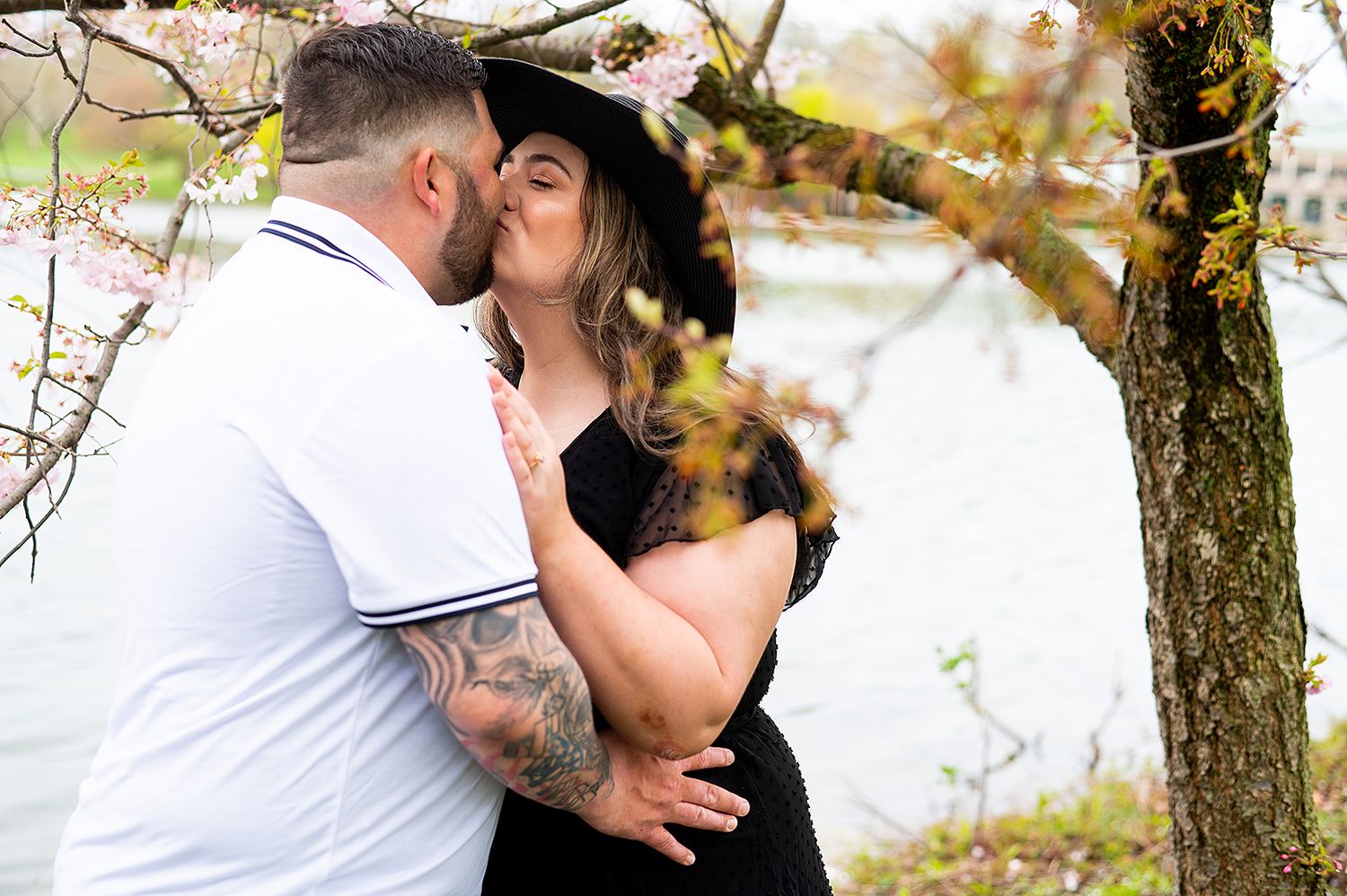 A romantic couple embraces while kissing under spring blossoms near a waterfront, with her wearing a black hat.
