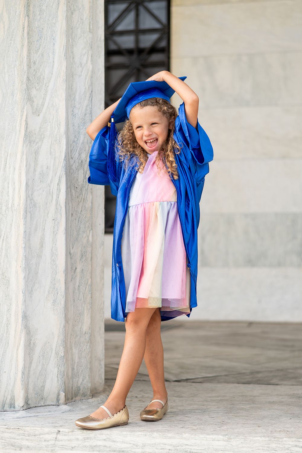 Young graduate in blue gown and pink dress poses playfully while holding graduation cap overhead.