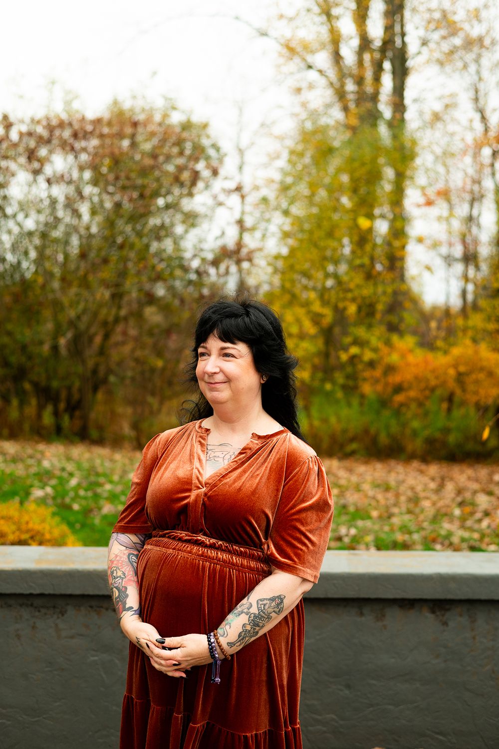 A woman in a rust-colored velvet dress poses outdoors against an autumn backdrop with changing leaves.