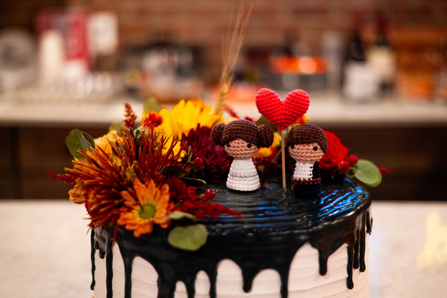 Chocolate drip cake decorated with red heart and small doll figures on top alongside orange and red fall flowers.