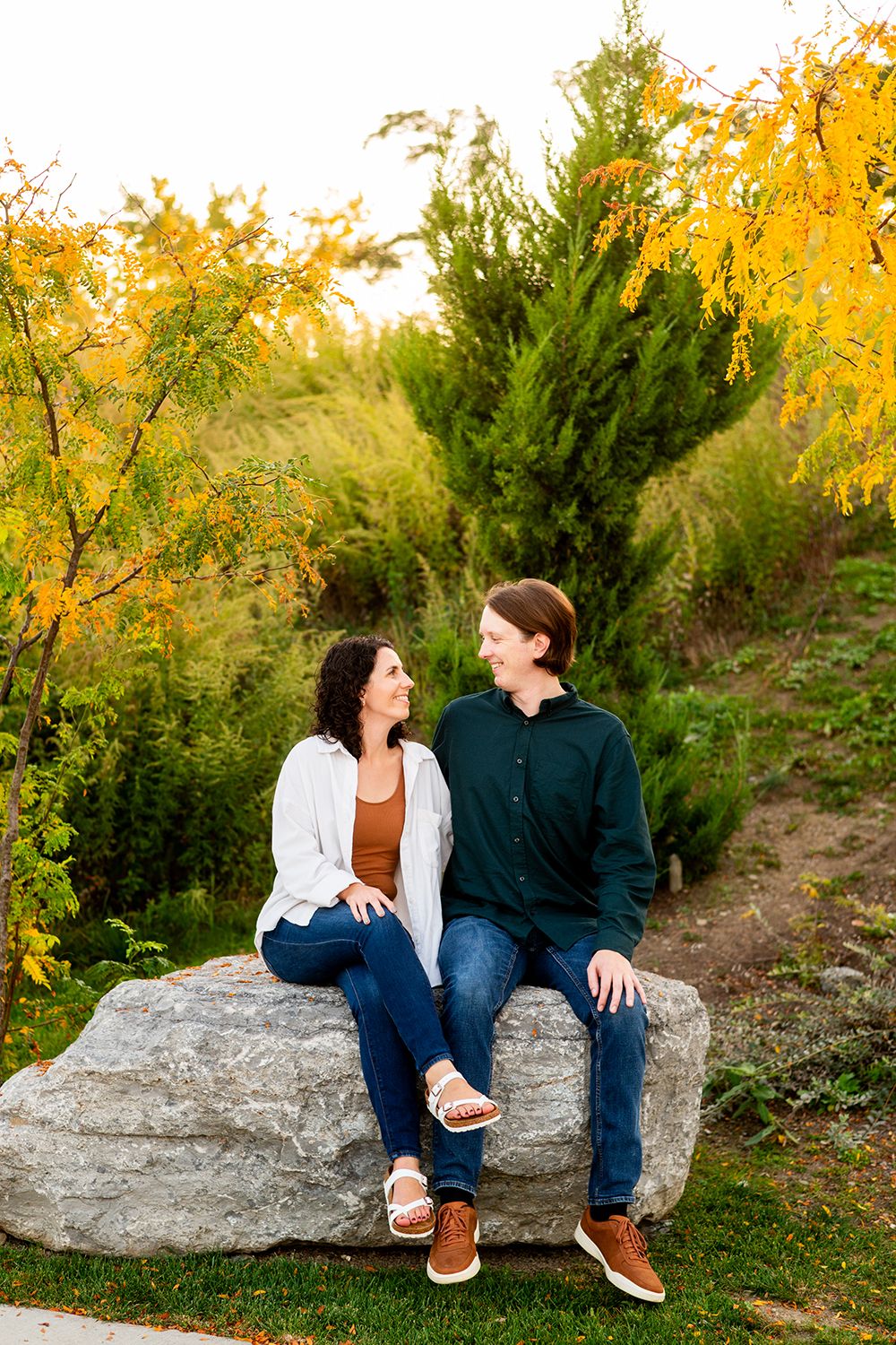 A happy couple sits on a large boulder during autumn with golden foliage behind them at sunset.