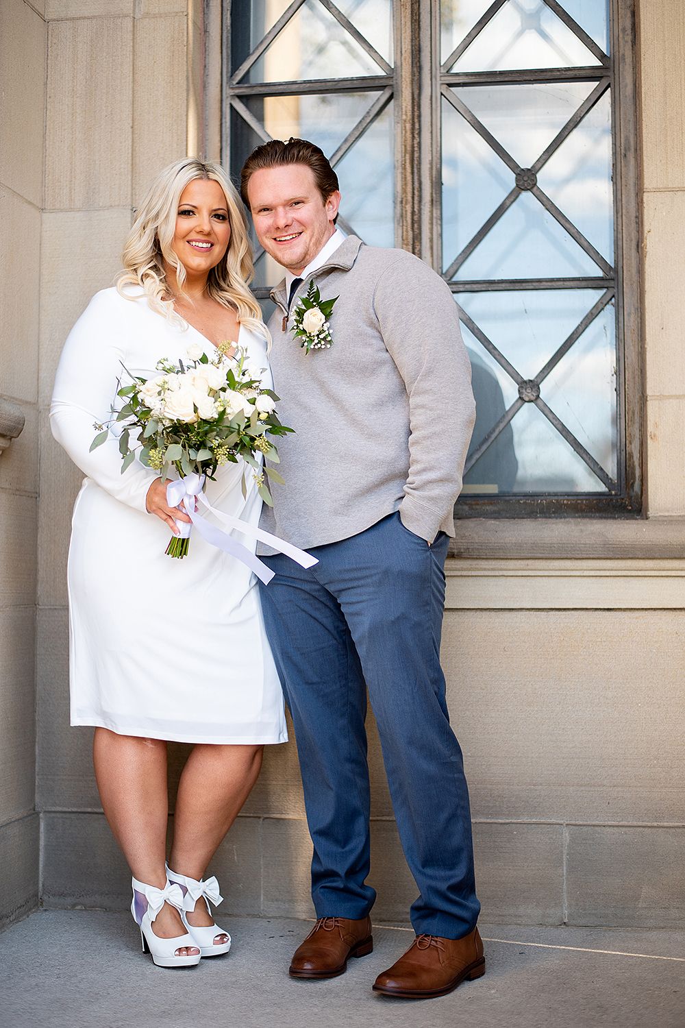 A couple poses in front of a decorative window at a historic building entrance wearing casual wedding attire.