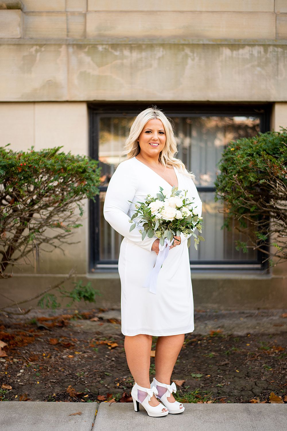 A bridal portrait outside a building showing a person in a short white dress holding a bouquet of white flowers.