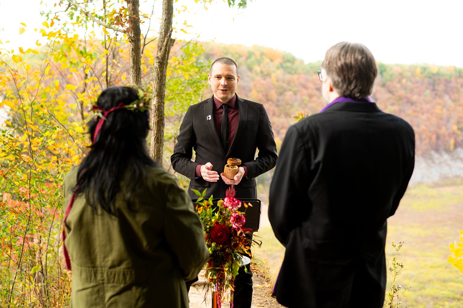 A small wedding ceremony takes place outdoors during autumn with fall foliage in the background.