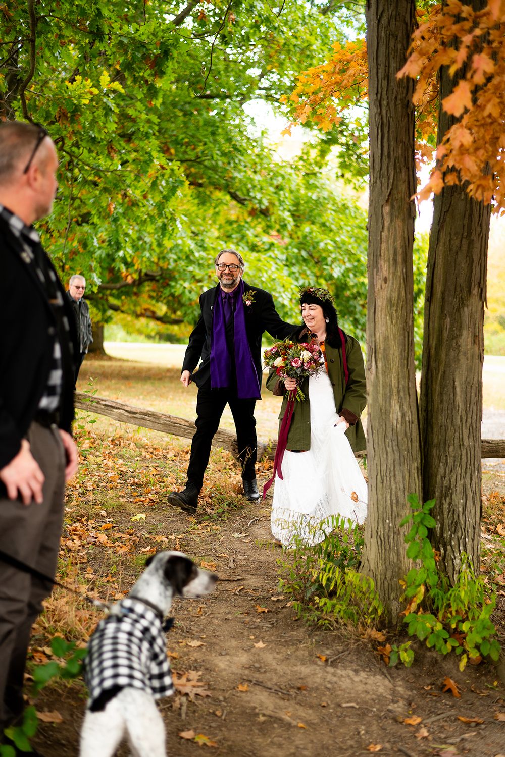 Small dog playfully runs between trees during an autumn outdoor gathering in a wooded area.