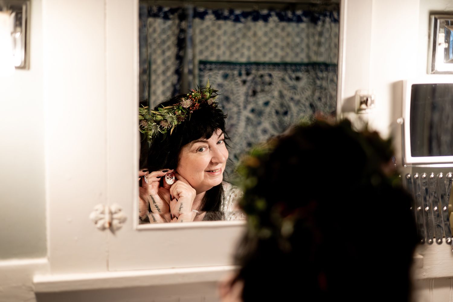 A person looks into a vintage-style bathroom mirror with floral pattern wallpaper and soft lighting.