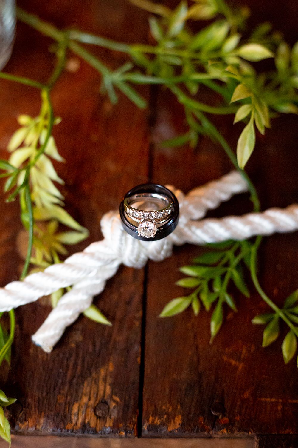 Diamond engagement ring balanced on white rope with green leaves on dark wooden background.