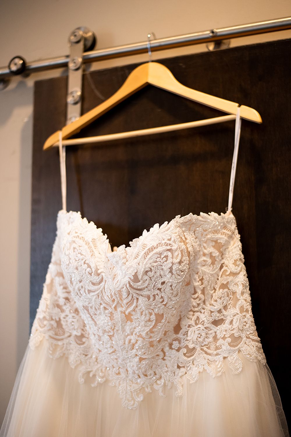A delicate white lace wedding dress with intricate floral patterns hangs on a wooden hanger against a dark wall.