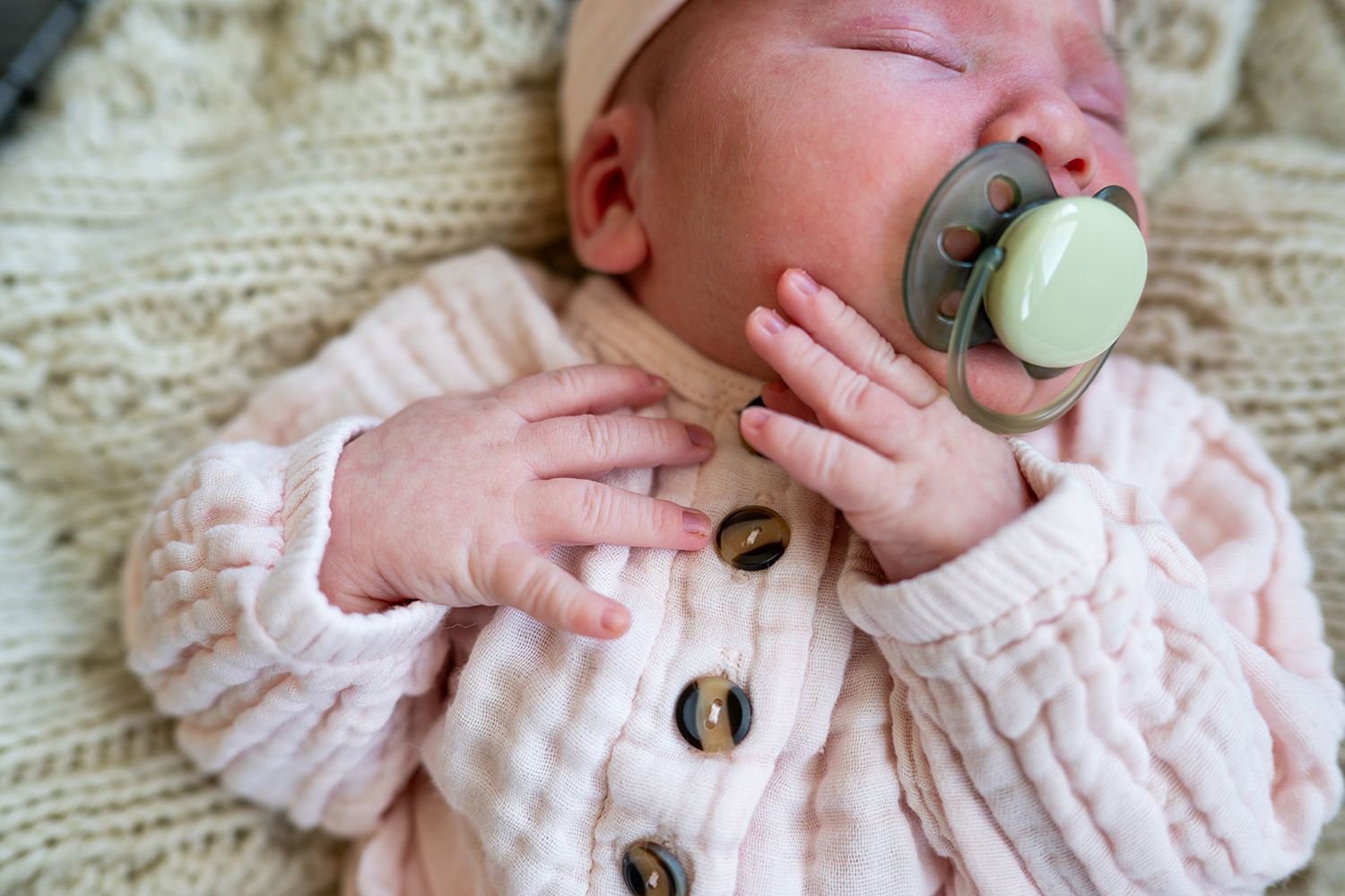 Close-up of newborn baby's hands wearing pale pink cardigan while holding pacifier on knit blanket.