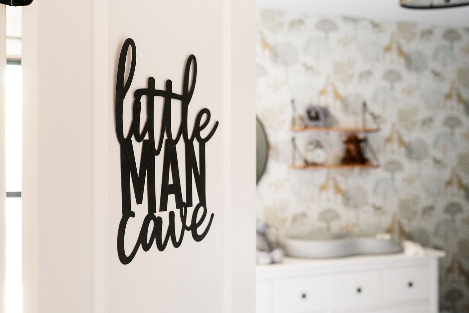 Modern black 'Little Man Cave' text decal mounted on white wall with nursery furniture in background.