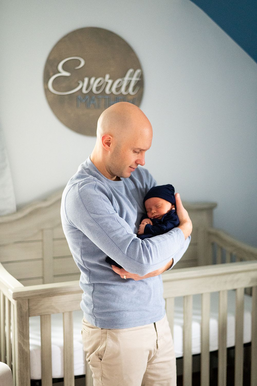 A tender nursery scene with a parent in gray sweater holding a sleeping newborn in front of a crib and name wall decor.