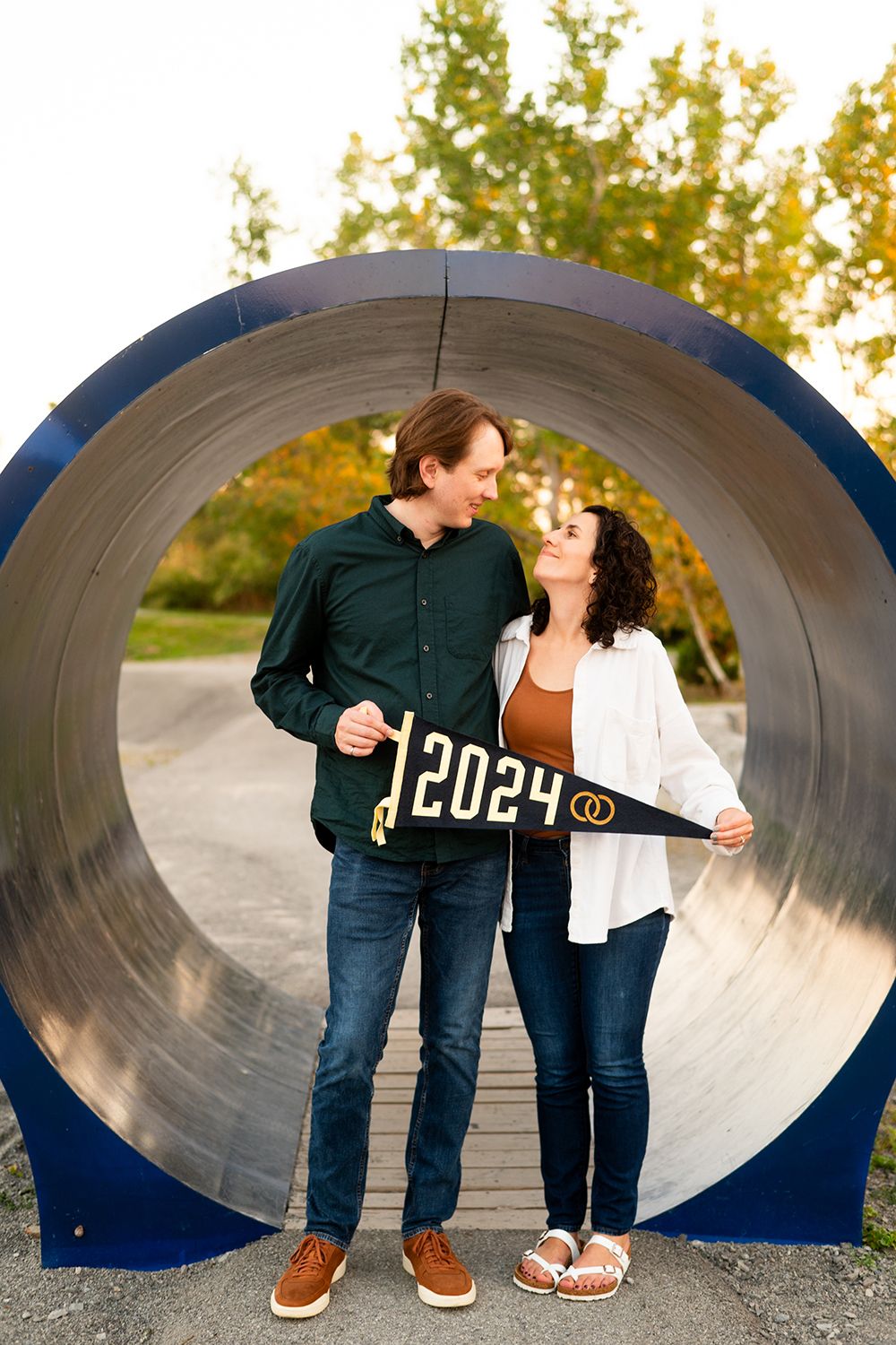 A couple poses inside a large circular tunnel while holding up 2024 numbers for an engagement photo.