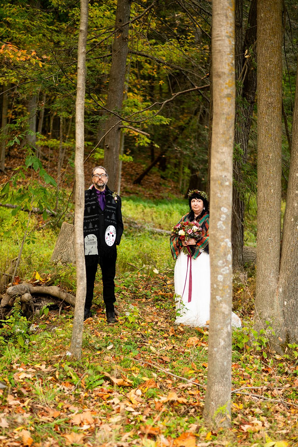 A couple pose in a woodland setting during fall, surrounded by tall trees and scattered autumn leaves on the ground.