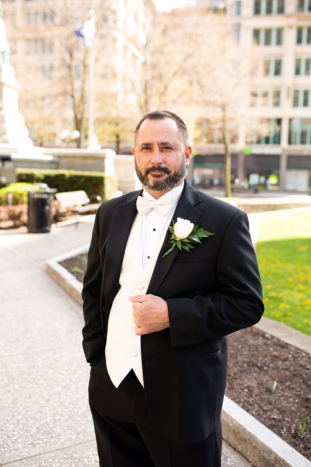 A groom in formal black tuxedo and white bow tie with boutonniere stands outside on an urban pathway.