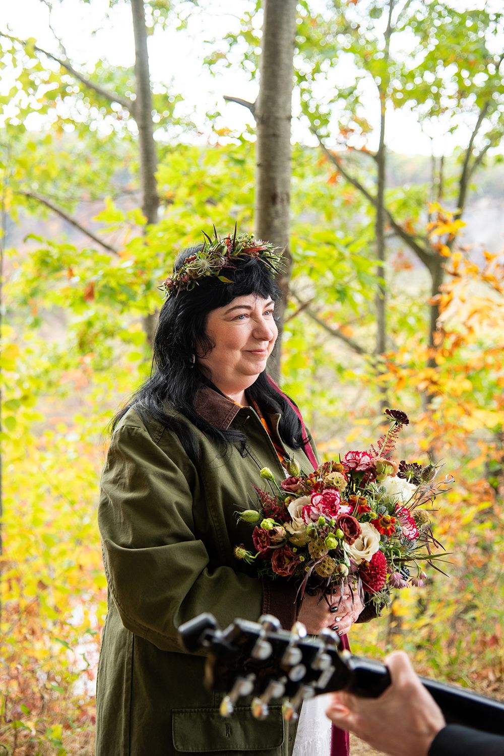 Person in green jacket smiles while holding autumn bouquet in wooded outdoor setting with fall foliage.