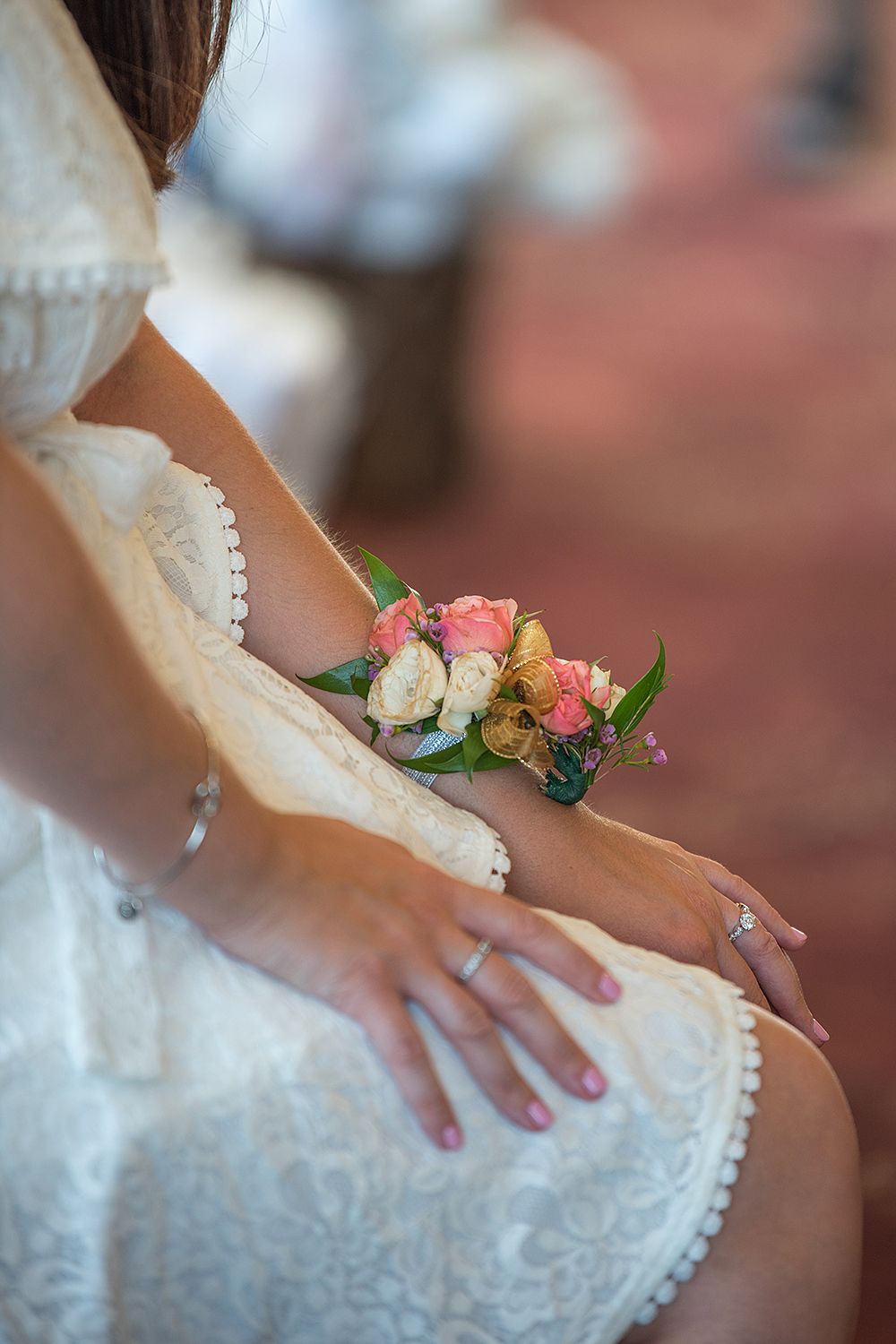 A delicate pink and white corsage adorns a bride's wrist against her lace wedding dress.
