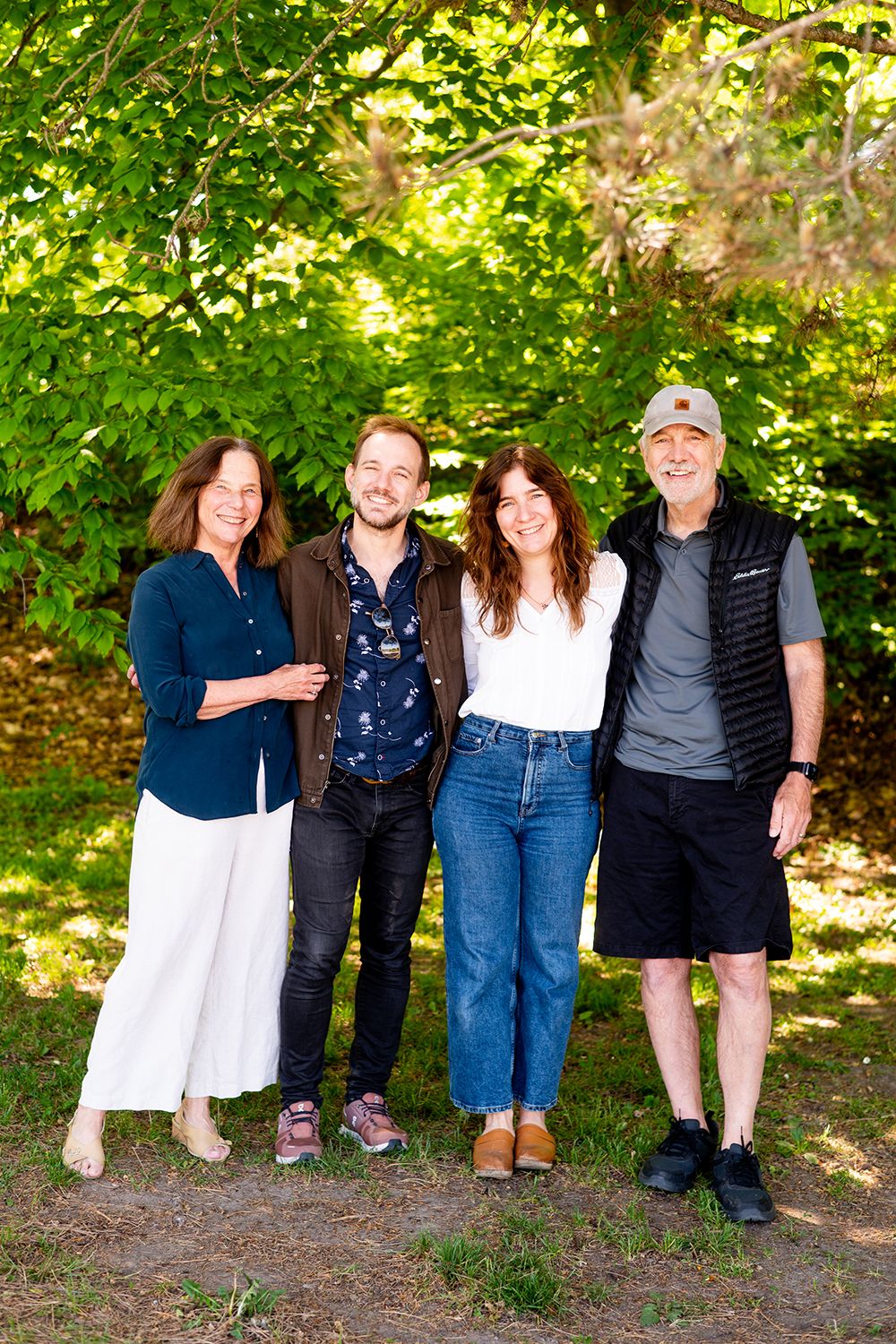 Casual family of four posing together outdoors under green leafy trees on a sunny day.