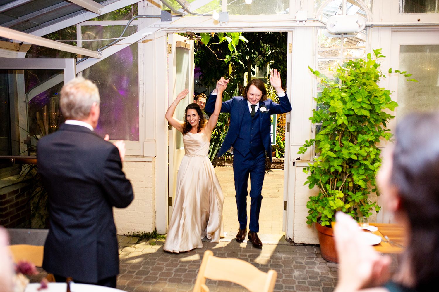 Wedding couple makes grand entrance through greenhouse doorway as guests celebrate.
